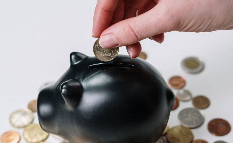 Hand inserting a coin into a black piggy bank with scattered coins around it.