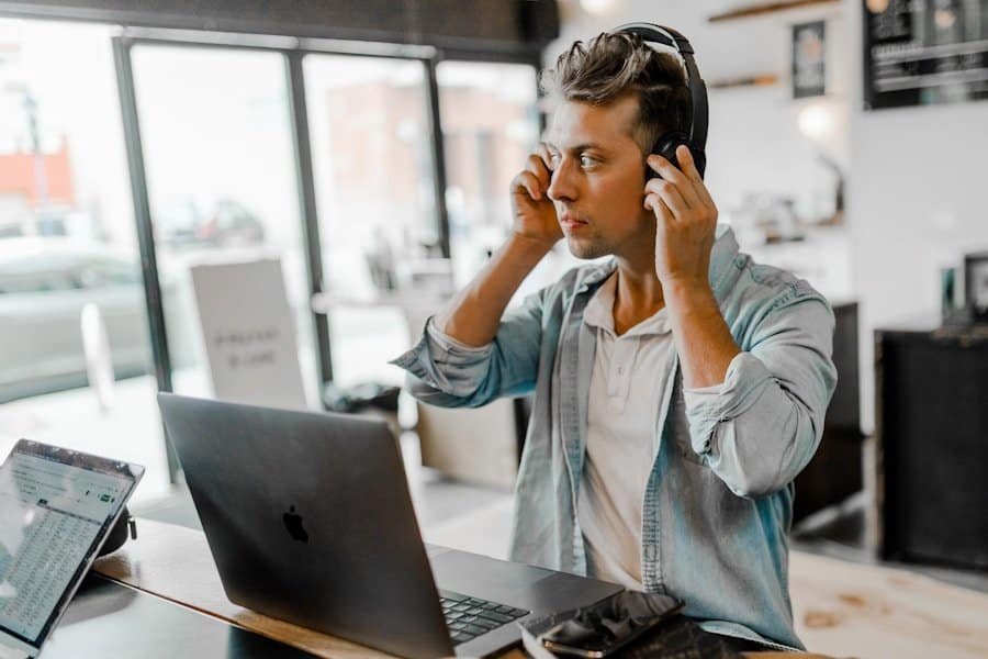 Man in a casual shirt using headphones while working on a laptop in a modern cafe.