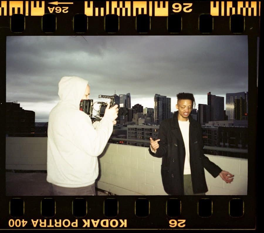 Person in a hoodie filming another person on a rooftop with a city skyline in the background.