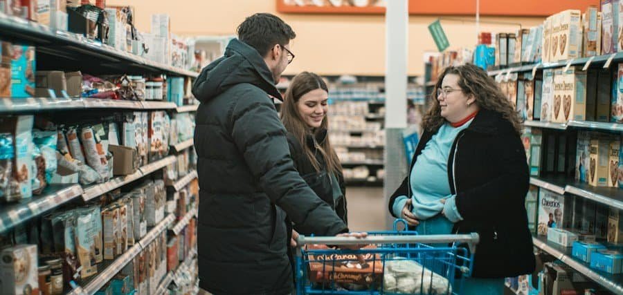 Three people conversing near a shopping cart in a grocery store aisle.