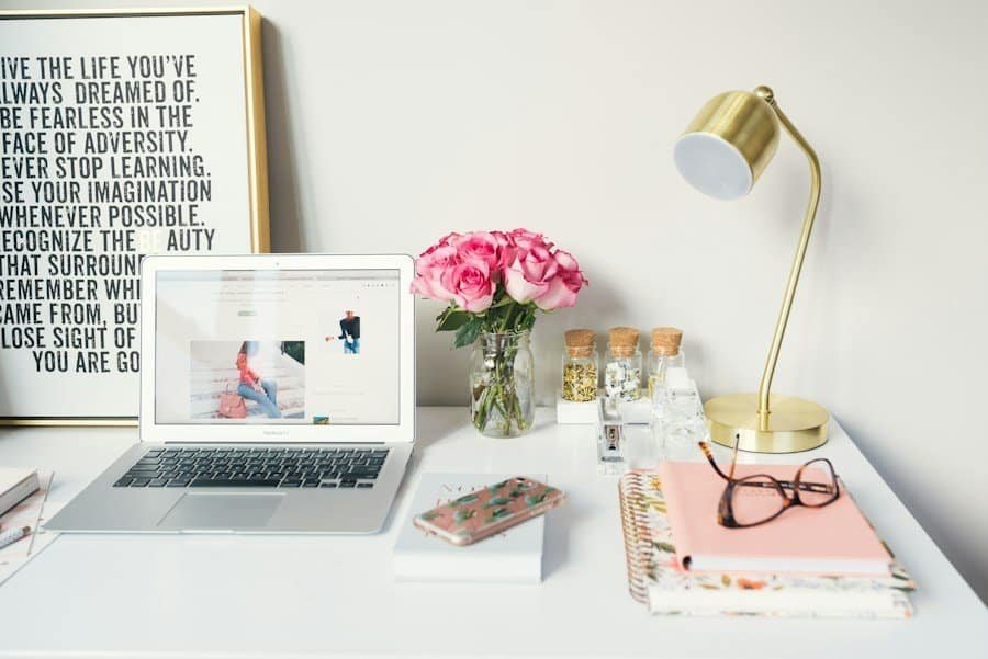 A modern workspace with an open laptop, a desk lamp, a bouquet of pink roses, framed inspirational quote, and stationary supplies.