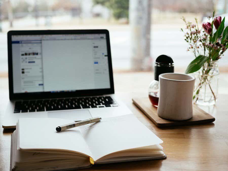 A laptop opened to a social media site next to an open notebook with a pen and a mug of tea on a wooden desk.