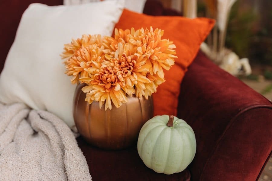 A cozy autumn-themed setting with orange flowers arranged in a pumpkin vase, accompanied by a green pumpkin and colorful cushions on a red chair.